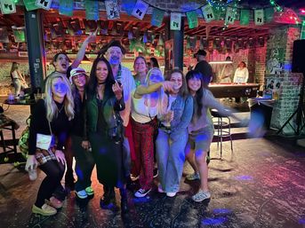 Group of friends enjoying a night out in a lively urban bar with exposed brick walls, hanging green banners and string lights, colorful disco lighting, and a pool table in the background.