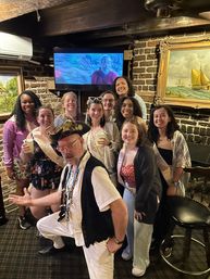 Group of smiling friends in a cozy brick-walled pub, holding drinks and wearing sashes, posing with a costumed pirate host under wooden beams and nautical artwork.