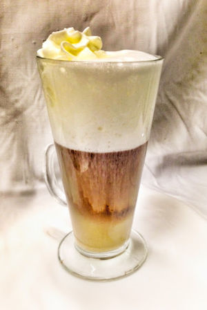 Tall layered latte macchiato in a footed clear glass — milk at the bottom, dark espresso center, thick frothy milk and a whipped cream swirl on top with steam rising against a soft white background.