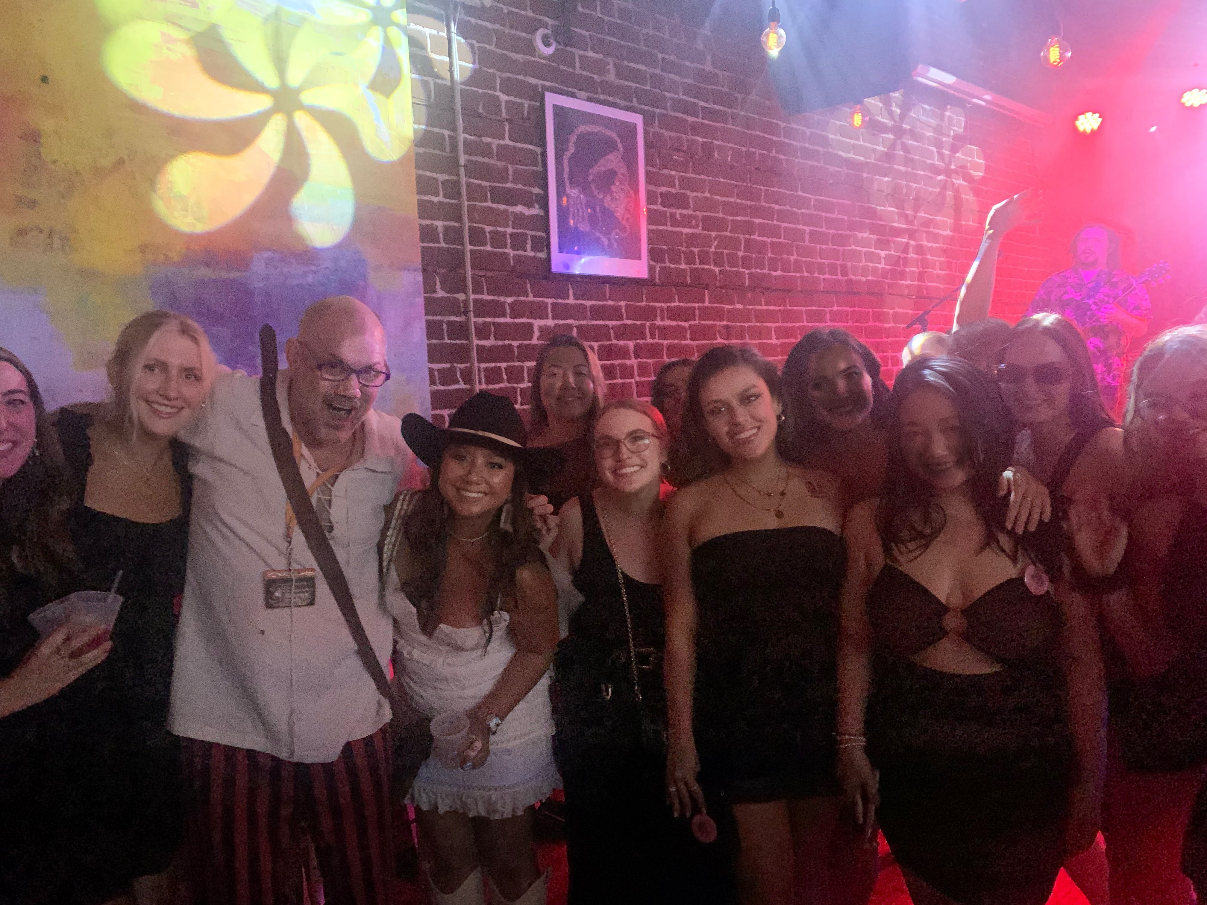 Smiling group of friends posing at a lively nightclub with exposed brick wall, colorful stage lights, live music, drinks and a person wearing a cowboy hat.