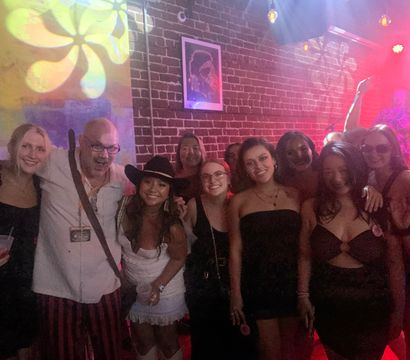Smiling group of friends posing at a lively nightclub with exposed brick wall, colorful stage lights, live music, drinks and a person wearing a cowboy hat.
