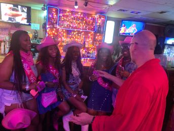Smiling women in pink cowboy hats and 'Bride Tribe' sashes gathered at a neon-lit bar with drinks while a man in red interacts — bachelorette night out