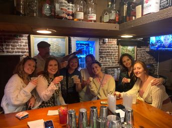 Group of friends toasting with shots at a cozy brick-walled bar — wooden countertop, cocktail shakers, bottles on a shelf, lively night out.