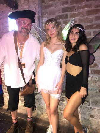 Three people posing against an exposed brick wall at a costume party: a man in a tricorn hat, white shirt and pink glasses, a woman in a white fairy dress with wings and floral crown, and a woman in a black cutout outfit with wings and jeweled headpiece.
