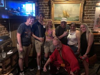 Group of seven people posing in a cozy brick-walled pub under a framed sailboat painting, holding drinks—one wearing a sash and tiara and another crouched in a red costume, casual summer outfits and warm bar lighting.