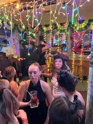Crowded neighborhood bar interior with colorful string lights and garlands, people dancing by a small stage and microphone; woman in a pirate hat making a surprised face, another holding a cocktail, beer sign and graffiti-covered walls in the background.