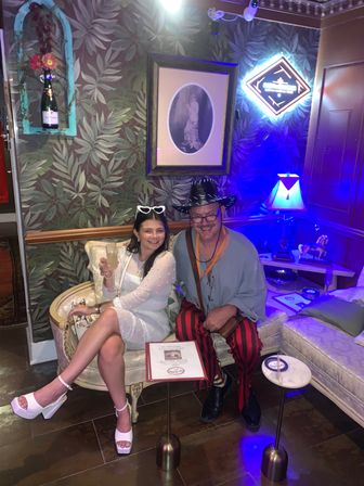 Smiling duo in a retro cocktail lounge on a velvet loveseat — woman in a white dress and heart sunglasses holding a cocktail, man in a black cowboy hat and red-striped pants under a neon sign, surrounded by leafy wallpaper, framed portrait and eclectic lamps.