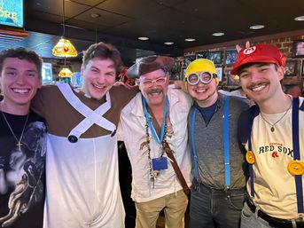 Five smiling people in costume pose arm‑in‑arm inside a cozy pub — pirate with tricorn hat, minion with yellow hat and goggles and blue suspenders, person in white overalls, person in red Mario cap wearing a Red Sox T‑shirt, stained‑glass lamps and brick wall background.