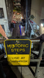 Worn yellow sign reading "HISTORIC STEPS – USE AT OWN RISK" on a rusty railing overlooking narrow wet city steps and an alley lit by hanging string lights at night.