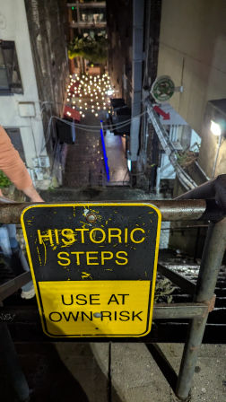 Worn yellow sign reading "HISTORIC STEPS – USE AT OWN RISK" on a rusty railing overlooking narrow wet city steps and an alley lit by hanging string lights at night.