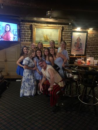 Smiling group of women in light blue dresses posing with a costumed man in red-striped pants inside a cozy brick-walled bar, nautical painting on the wall and drinks on a high table.