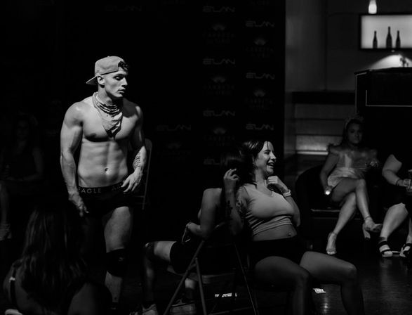 Black-and-white photo of a shirtless male dancer in a backwards cap performing beside seated, laughing women in a dim nightclub setting.