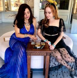 Two women in blue and black evening gowns seated on a white ottoman in a boutique-style room, reading three tarot cards on a small carved wooden table with a candle and crystals, wearing statement jewelry and a yellow headpiece.