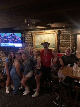 Group of people posing in a dim, historic brick pub with exposed wooden beams, a framed sailboat painting and a wall TV showing sports — one person in a playful tricorn hat, others in casual outfits, drinks on a wooden table for a lively night out.