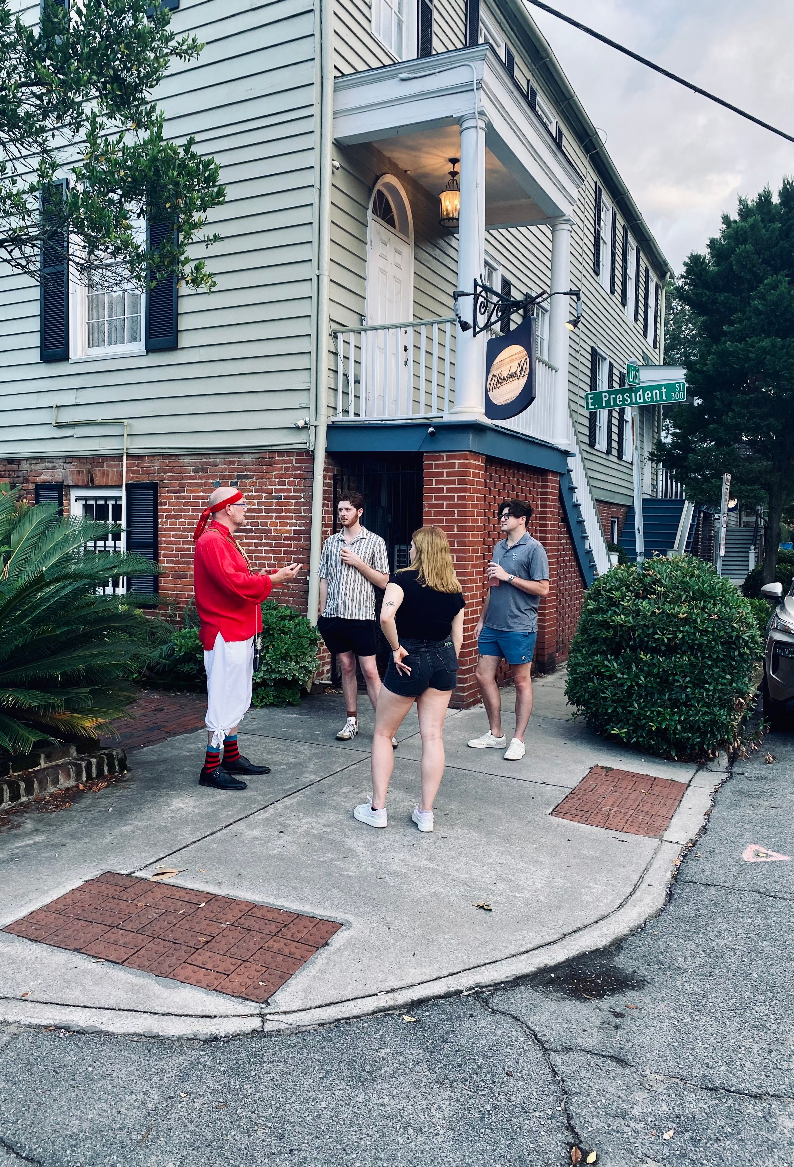 Four people chatting on a street corner outside a clapboard-and-brick historic house with white porch columns and an 'E. President St' sign