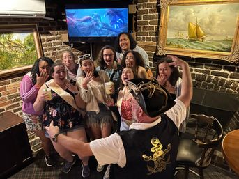 Excited group of friends at a bachelorette party in a cozy brick-walled bar, gathered around a pirate-hatted entertainer with outstretched arms; women wearing sashes hold cocktails beneath framed nautical paintings and a TV showing a sea scene.