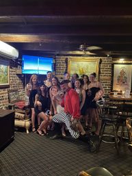 Group of women in black dresses posing with a costumed pirate entertainer kneeling in front, holding drinks and paper face cutouts inside a cozy brick‑walled pub with nautical paintings and bar stools.