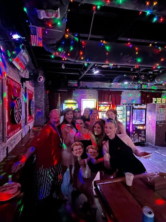 Group of friends posing and smiling in a dim, arcade-style bar decorated with multicolored string lights, neon game machines, dartboards on exposed brick walls, and drinks on a wooden table — lively nightlife scene.