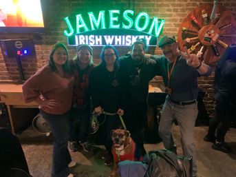 Five people smiling in a dim brick-walled pub beneath a glowing green neon whiskey sign, posing together with a leashed brown boxer dog wearing a red sweater.