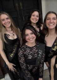 Four smiling women in black dresses wearing celebratory sashes, posing together in front of a dark double door — group portrait of friends at an evening event.