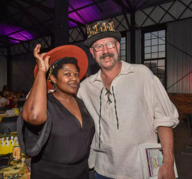 Two smiling attendees in an industrial event space — a Black woman tipping an orange wide-brim hat and a man in a decorative top hat and linen shirt, purple uplighting and corrugated metal walls in the background.