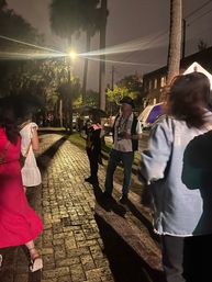 Rainy nighttime scene on a palm-lined brick sidewalk in a downtown area, wet cobblestones glint under a bright streetlamp as people with umbrellas listen to a hat-wearing guide gesturing to the group.