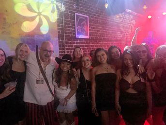 Smiling group of friends posing in an exposed-brick indoor music venue with colorful projected patterns and red stage lights as a live band plays, capturing a lively night-out atmosphere.