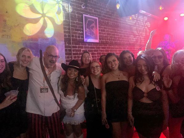 Smiling group of friends posing in an exposed-brick indoor music venue with colorful projected patterns and red stage lights as a live band plays, capturing a lively night-out atmosphere.