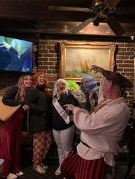 Laughing group in colorful wigs and costumes inside a cozy brick-walled pub, posing under a framed sailboat painting while a costumed pirate theatrically gestures.