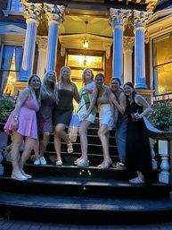 Seven friends posing on black front steps of a Victorian-style townhouse at dusk, lit Corinthian columns and stained-glass doorway in the background; one woman wearing a celebratory sash, summer dresses and casual shoes, some holding drinks.