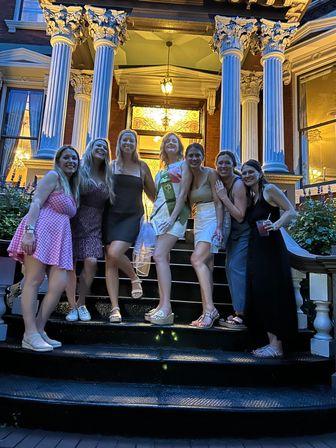 Seven friends posing on black front steps of a Victorian-style townhouse at dusk, lit Corinthian columns and stained-glass doorway in the background; one woman wearing a celebratory sash, summer dresses and casual shoes, some holding drinks.