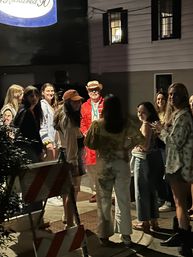 Nighttime sidewalk scene in a small-town downtown: a group of smiling young adults gathered around a man in a red coat and straw hat under warm streetlights, with an orange construction barrier in the foreground.