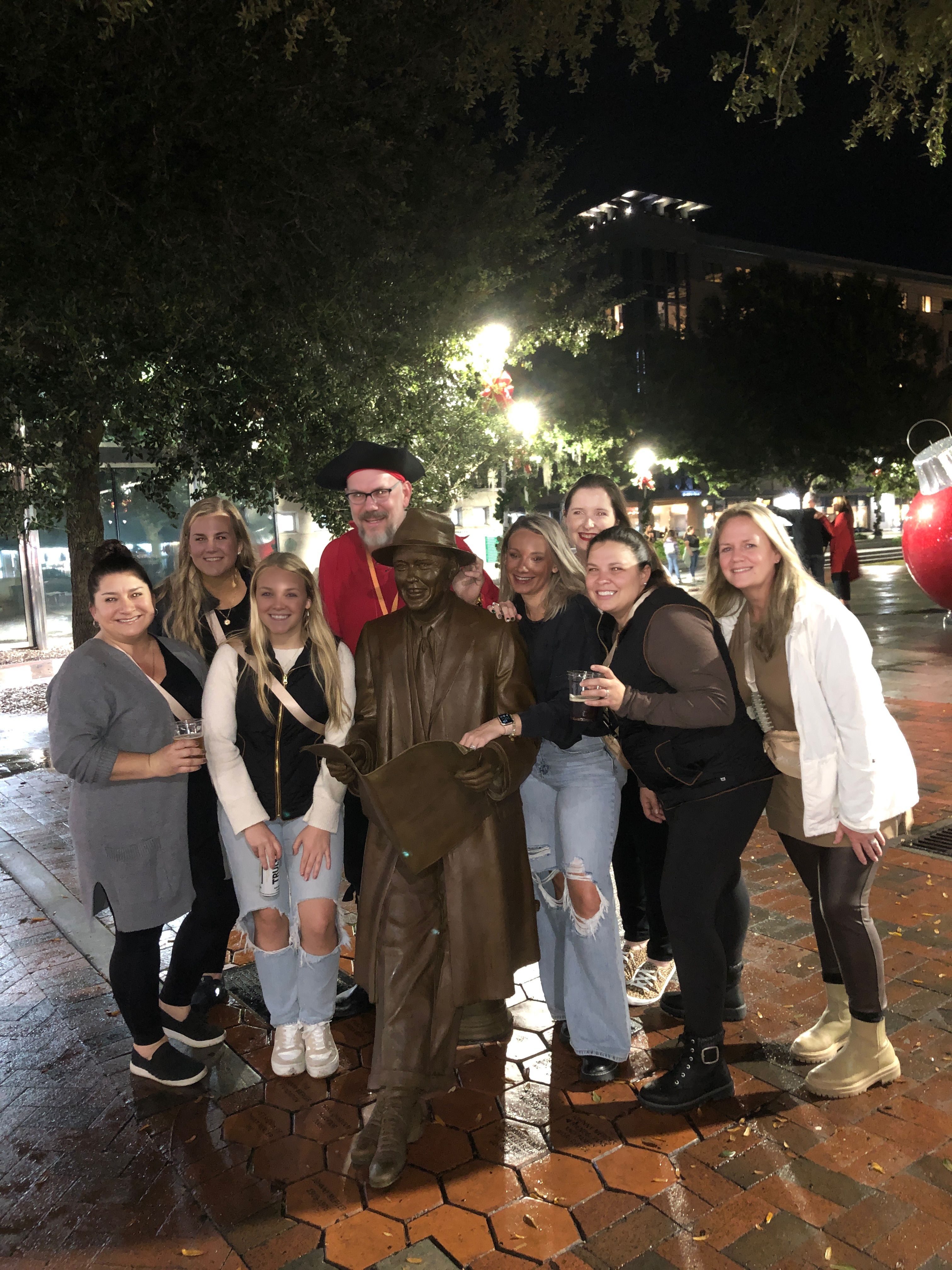 Cheerful group of friends posing at night around a bronze statue of a man holding a newspaper in a lit downtown plaza with wet brick pavement and holiday lights.