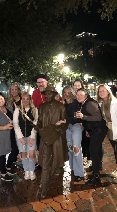 Group of smiling adults posing around a bronze statue of a seated man reading a newspaper in a lit downtown plaza at night, wet brick pavement and holiday decorations visible.