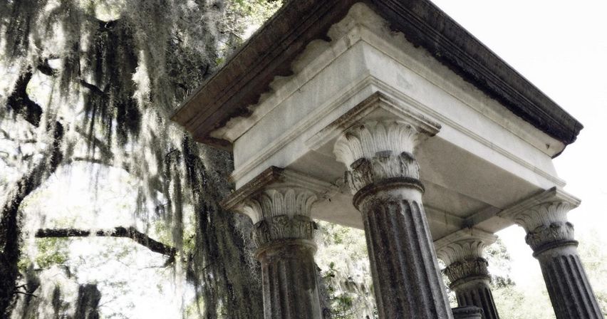 Weathered classical stone columns and entablature beneath Spanish-moss-draped trees, sunlit historic garden structure.