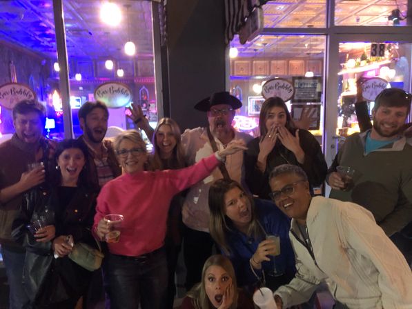 Group of friends laughing and posing outside a neon-lit bar at night, holding drinks and making surprised, playful faces.