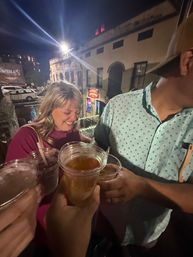 Friends cheerfully clinking plastic cups of beer on a rooftop balcony overlooking a historic downtown nightlife street at night