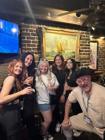 Group photo of friends on a night out in a cozy brick-walled pub — five women with drinks and a man in a tricorn hat posing in front of a framed sailboat painting.