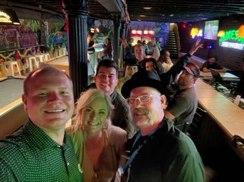 Group selfie of smiling friends in a neon-lit neighborhood bar with colorful mural art, shuffleboard lanes and a long wooden bar — lively nighttime scene.