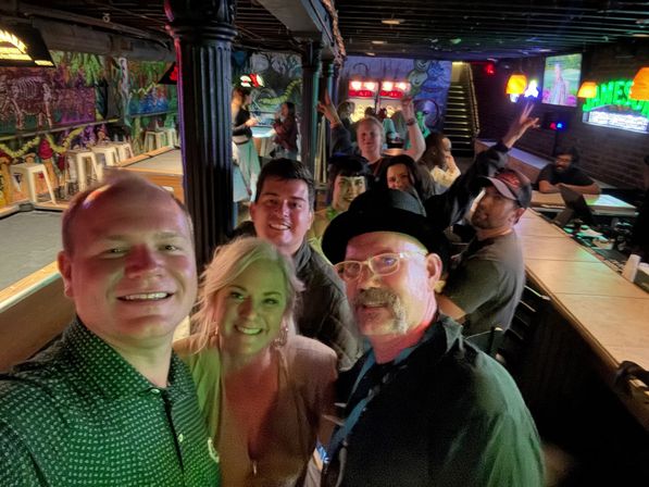 Group selfie of smiling friends in a neon-lit neighborhood bar with colorful mural art, shuffleboard lanes and a long wooden bar — lively nighttime scene.