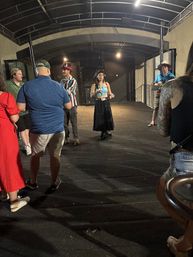 Evening crowd on a covered outdoor patio stage — a woman in a blue crop top and long black skirt stands center while casually dressed friends in hats chat nearby, a tattooed arm visible in the foreground.