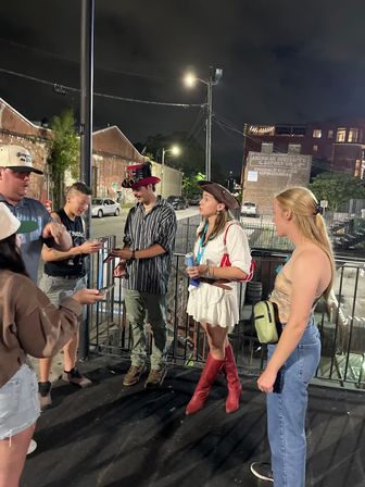 Group of young adults chatting on a downtown sidewalk at night by a metal railing — a woman in a white dress, red knee-high boots and a pirate hat holds a drink while a man in a striped shirt and steampunk top hat checks his phone, with brick buildings and streetlights in the background.