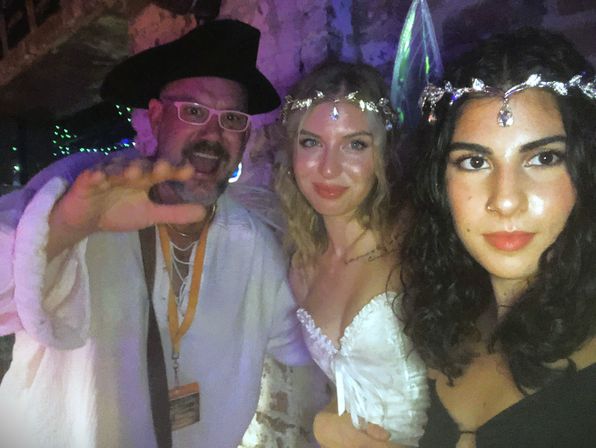 Selfie of three costumed partygoers in dim colorful lighting — a man in a white robe and hat reaching toward the camera and two women wearing jeweled headbands and sparkly outfits.