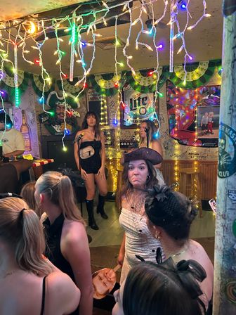 Festive pub interior with colorful string lights, women gathered for karaoke, singers on a small stage and a pirate-hat partygoer making a playful face.