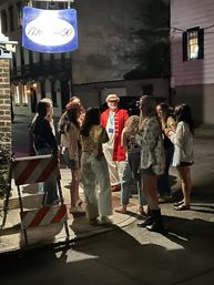 Nighttime downtown street scene: a group of young people gathered on a historic sidewalk around a man in a red vintage coat and straw hat with a lanyard, illuminated by warm streetlights beneath a hanging sign.