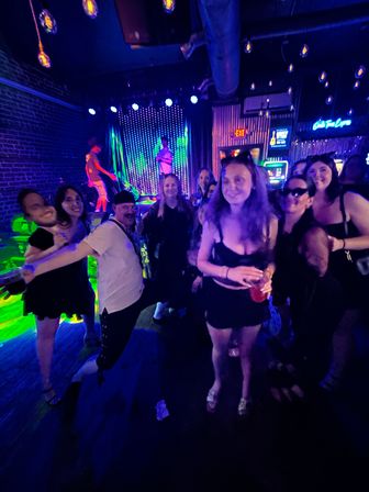 Neon-lit nightclub group photo of friends on a crowded dance floor with a live band on stage, purple and green lighting, exposed brick wall and hanging Edison bulbs, woman holding a red drink.