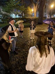 Nighttime downtown park scene: small group of adults standing in a circle under lamplight and trees, some holding umbrellas and wearing costumes — including a man in a decorated top hat — on a wet, leaf-strewn lawn.