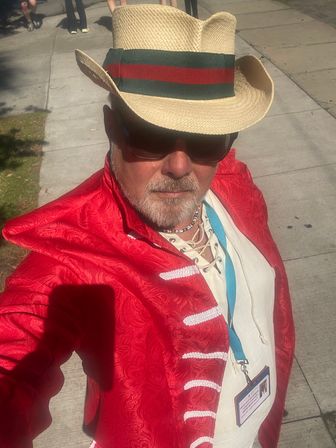Selfie of a person on a sunny urban sidewalk wearing a straw cowboy hat with a green-and-red band, dark sunglasses, white lace-up shirt, bright red brocade jacket and a blue lanyard badge.