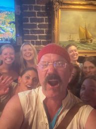 Cheerful group selfie inside a cozy brick-walled pub, man in a red beanie and glasses in front with smiling friends behind and a framed sailing painting on the wall.