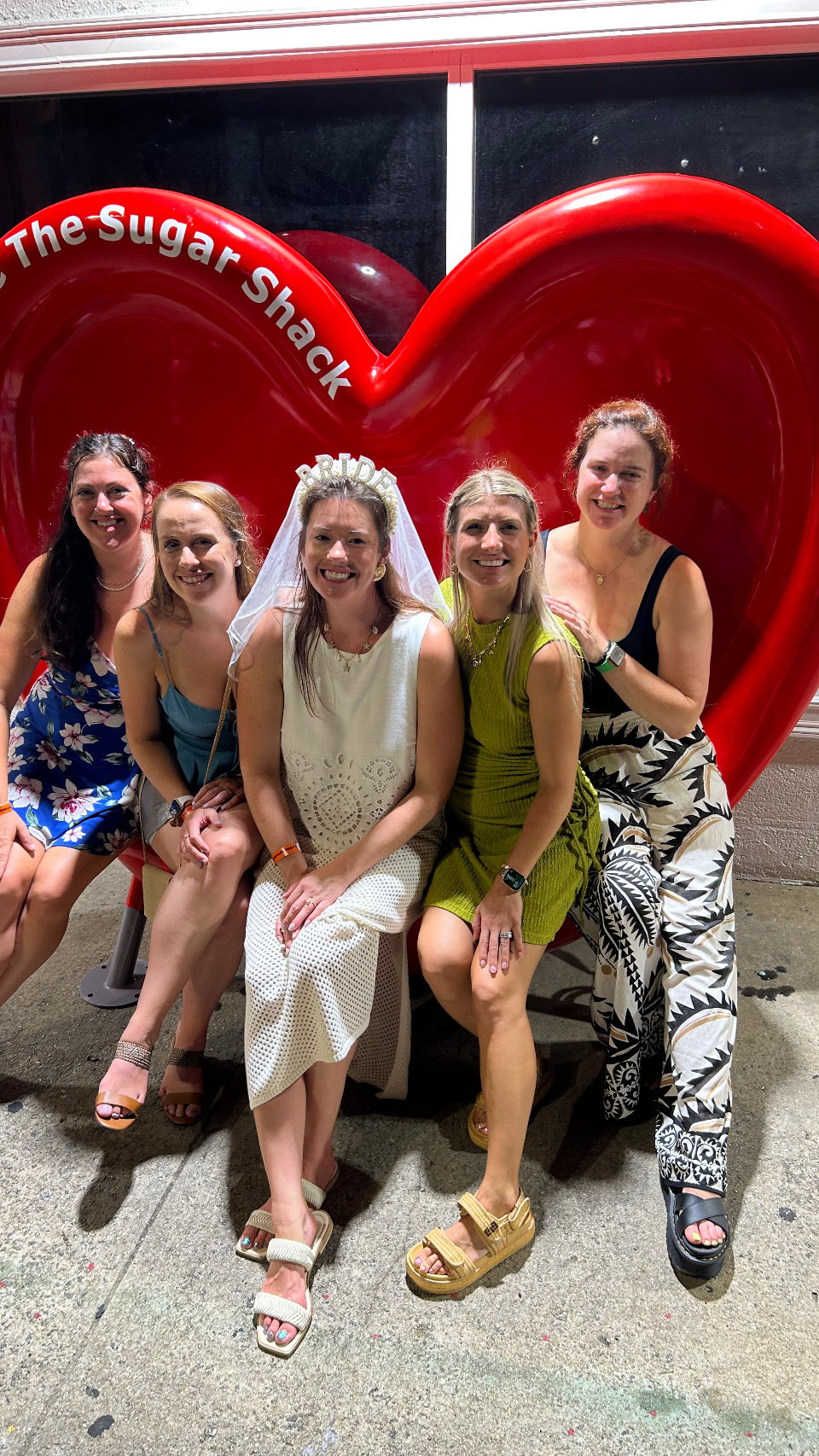 Laughing bride-to-be in a veil with four friends seated on an oversized red heart bench on a city sidewalk at night — fun bachelorette night out group photo.